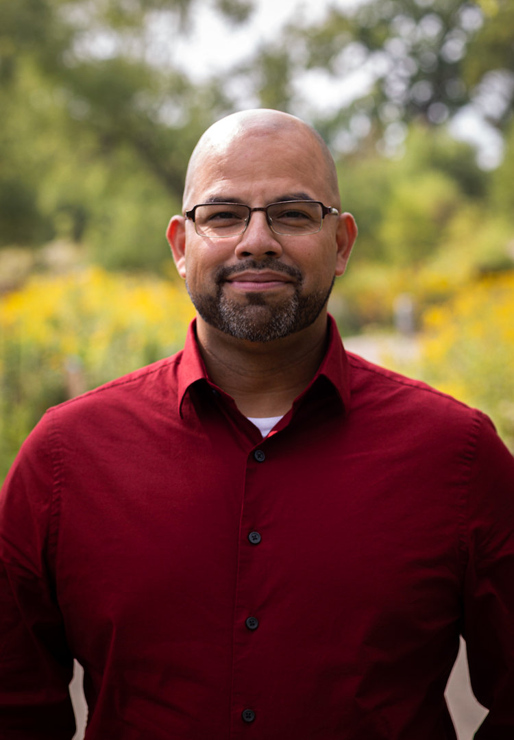 A headshot of Alderperson Andre Vasquez from the shoulders up. Wearing a red button up shirt, he is a 40something Latinx man with a bald head, glasses, a salt and pepper beard, and a warm smile.