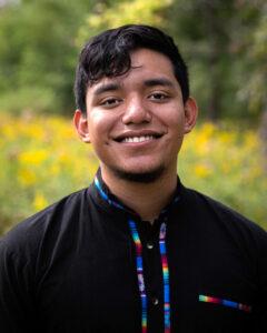 A headshot of Angel Rubi Navarijo from the shoulders up wearing a black button down shirt with colorful patterns on the seams where the buttons are aligned. He is a young man from Guatemala in his 20s with black hair, a gentle smile, and beautiful green trees around him.
