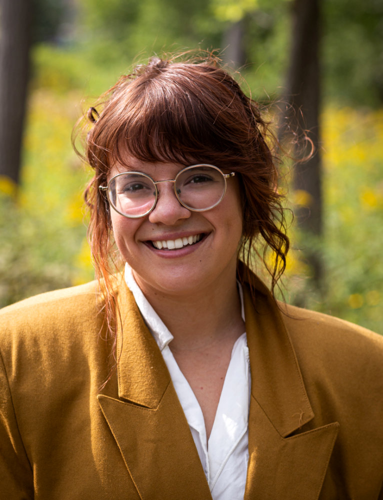 Morgan Madderom is wearing gold glasses, a mustard colored blazer and white colored shirt and has a joyful smile on her face. She is a 5'3" white woman with reddish brown wavy hair and long bangs.