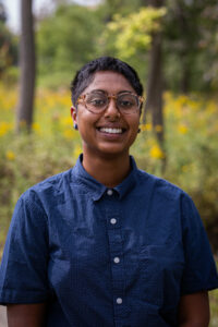 Pooja Ravindran is wearing glasses, a blue button down and smiling handsomely in front of yellow flowers. They are 5"5 nonbinary person of Indian descent, currently with close cropped black hair. 